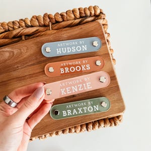 K&ouml;nnte beinhalten: Vier farbige Namensschilder aus Acryl mit dem Text "Artwork by" und einem Namen: Hudson, Brooks, Kenzie und Braxton. Die Schilder sind in Blau, Orange, Rosa und Gr&uuml;n gehalten und mit silberner Hardware befestigt. Die Schilder befinden sich auf einer Holzoberfl&auml;che.