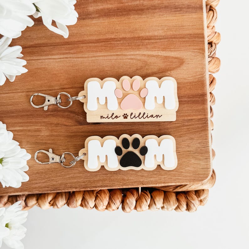 Puede incluir: Dos llaveros de madera con la palabra "MOM" en letras blancas. Un llavero tiene una huella de pata rosa, el otro una huella de pata negra. Los nombres "milo lillian" est&aacute;n grabados en el primer llavero. Ambos tienen llaveros plateados.