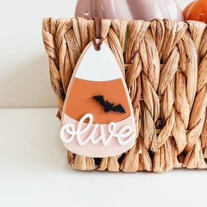 May include: A white, orange, and pink candy corn-shaped ornament with a black bat and the word "olive" written in white. The ornament is hanging from a wicker basket.