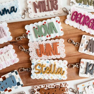 May include: A collection of rectangular name tags with scalloped edges, each featuring a unique patterned background and raised 3D lettering in various colours. Each tag includes a silver metal lobster clasp attached to one side.