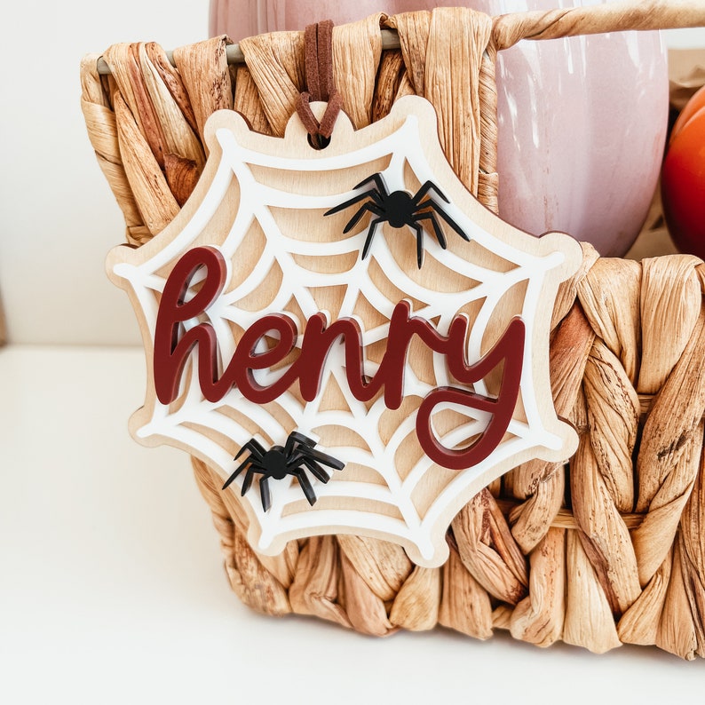 May include: A white wooden spiderweb cutout with the name "Henry" written in maroon letters. There are three black spider cutouts on the web.