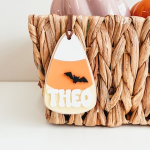May include: A white, orange, and yellow candy corn shaped ornament with a black bat and the name "THEO" written in white letters.