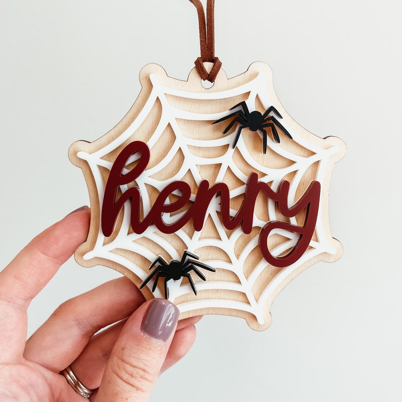 May include: A wooden spiderweb ornament with the name "Henry" written in red. The spiderweb is decorated with three black spiders.