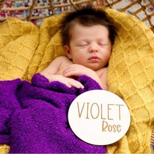 May include: A white wooden circle with the name "VIOLET Rose" written in gold lettering. The circle is lying on a purple blanket and a yellow knitted blanket. A baby is sleeping on the blankets.