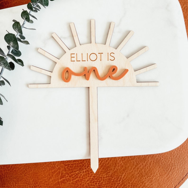 Puede incluir: Topper de pastel de madera con un dise&ntilde;o de sol y el texto "Elliot is one" en naranja.