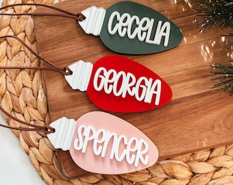 May include: Three lightbulb-shaped Christmas ornaments with names "Cecelia", "Georgia", and "Spencer" written in white on them. The ornaments are green, red, and pink.