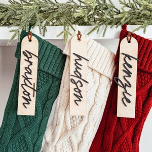 Peut inclure: Trois chaussettes de Noël avec des étiquettes en bois personnalisées. Les chaussettes sont vertes, blanches et rouges avec un motif en tricot torsadé. Les étiquettes sont brunes avec des lettres noires. Les étiquettes indiquent "Braxton", "Hudson" et "Kenzie".