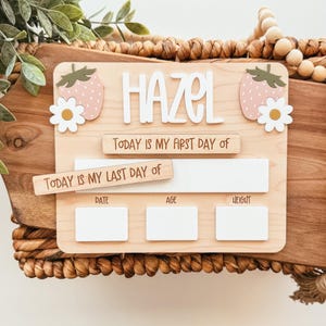 May include: Wooden milestone board with the name "HAZEL" in white letters. The board features strawberry and daisy accents, with spaces for "DATE", "AGE", and "HEIGHT". Includes the phrases "TODAY IS MY FIRST DAY OF" and "TODAY IS MY LAST DAY OF".
