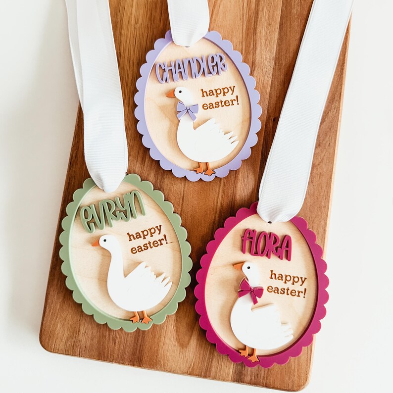 May include: Three wooden Easter egg ornaments with scalloped edges. Each ornament features a white goose with a pink bow, a name, and the text "happy easter!" The ornaments are in shades of green, purple, and burgundy.