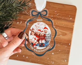 May include: A clear acrylic Christmas ornament with a scalloped edge and a dark blue border. The ornament features a photo of a family wearing red and white holiday clothing, the text 2025, and the text THE WILSON FAMILY, topped with a white ribbon.