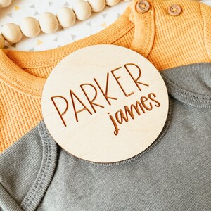 May include: A round wooden sign with the name "Parker James" engraved in a cursive font. The sign is on top of a gray and yellow baby onesie.