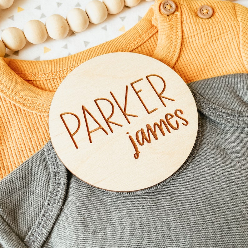 May include: A round wooden sign with the name "Parker James" engraved in a cursive font. The sign is on top of a gray and yellow baby onesie.
