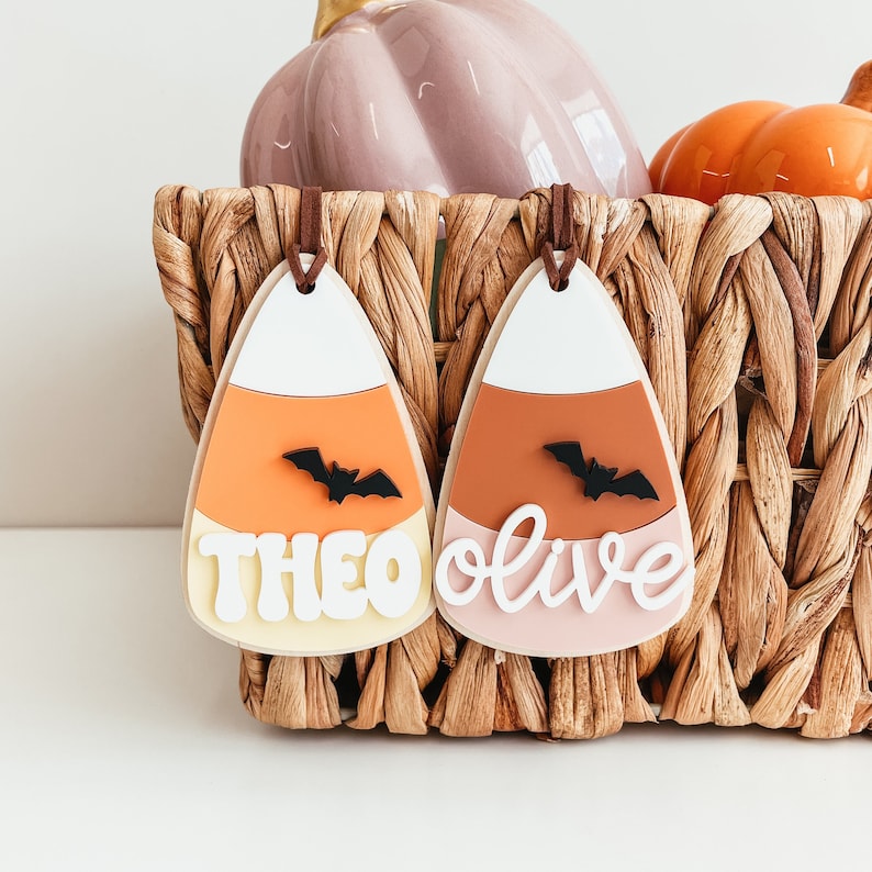May include: Two candy corn shaped ornaments with the names Theo and Olive written on them. Each ornament has a black bat silhouette on the orange section.
