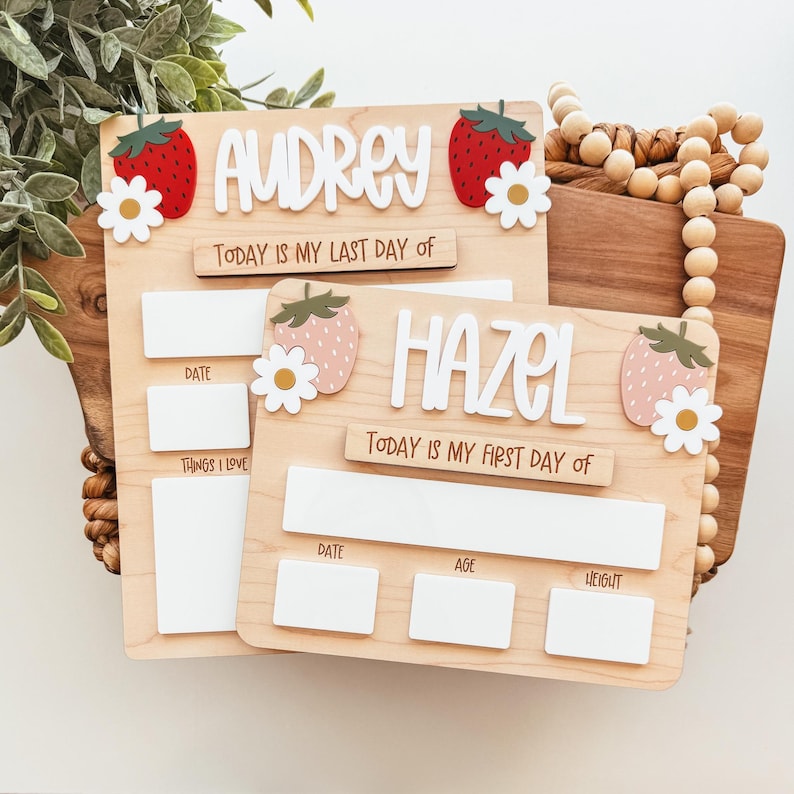 May include: Two wooden milestone boards with the names "Audrey" and "Hazel" in white letters. Each board features strawberry and daisy accents. Text includes "Today is my last day of" and "Today is my first day of".