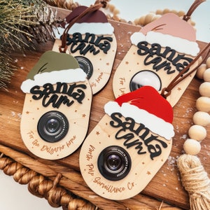 May include: Four wooden ornaments shaped like Santa hats with a camera lens in the center. Each ornament has the text "Santa Cam" and a different colored Santa hat. The text "Lens Made in Santa's Workshop" is printed on the ornaments.