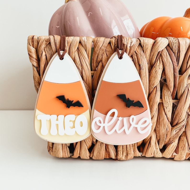 May include: Two candy corn shaped ornaments with the names Theo and Olive written on them. Each ornament has a black bat on it.