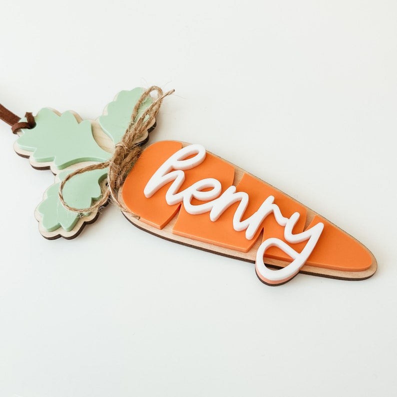 May include: A wooden carrot-shaped ornament with a green leafy top and the name "Henry" written in white on the orange carrot.