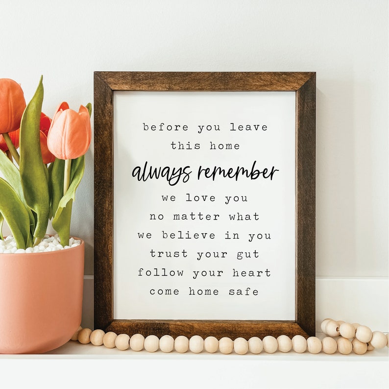 May include: A framed print with a white background and black text that reads "before you leave this home always remember we love you no matter what we believe in you trust your gut follow your heart come home safe". The frame is brown wood.