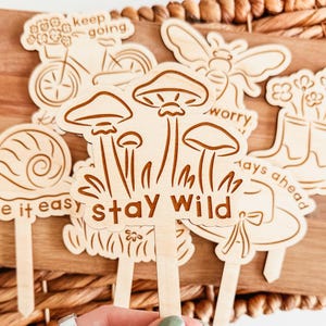 May include: A collection of wooden garden stakes featuring engraved designs of a bicycle, snail, bee, and mushrooms. The mushroom stake has the words "Stay Wild." Other stakes have the phrases "Keep Going," "Be it easy," and "Worry Less."