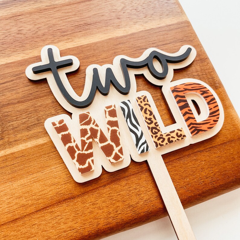 Peut inclure: Topper de g&acirc;teau "Two Wild" avec une inscription en &eacute;criture cursive noire et des lettres &agrave; imprim&eacute; animal. Les lettres sont en bois et ont une palette de couleurs marron, orange et noir.