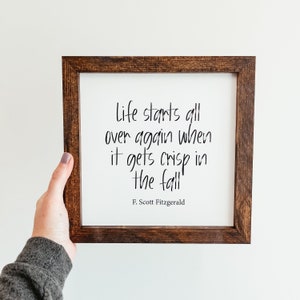 May include: A framed sign with a white background and black text that reads "Life starts all over again when it gets crisp in the fall" by F. Scott Fitzgerald. The frame is a dark brown colour.