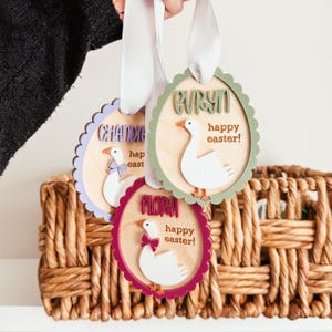 May include: Three wooden Easter ornaments shaped like geese with scalloped edges. Each ornament has a different name, "Flora", "Evelyn", and "Chandler", and the text "Happy Easter!"  The ornaments are painted in different colors: purple, green, and red.