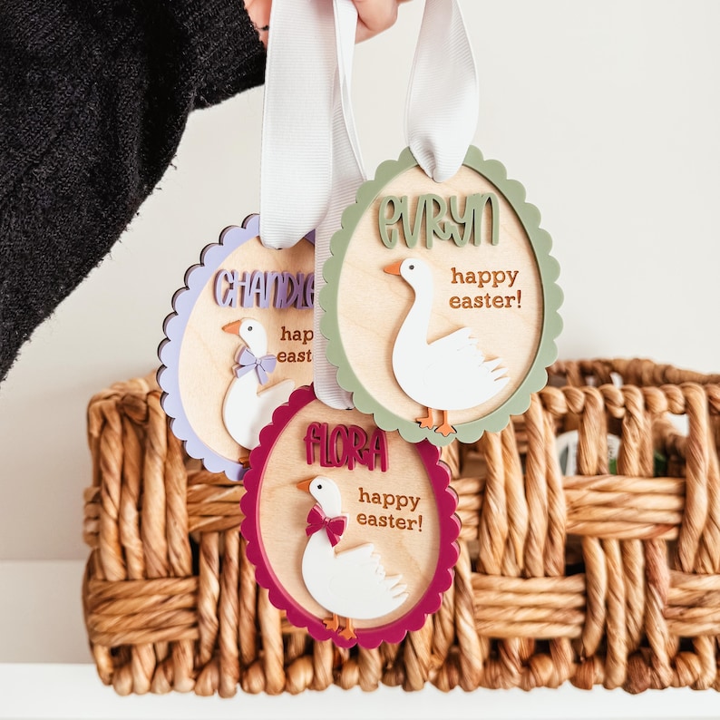 May include: Three wooden Easter ornaments shaped like geese with scalloped edges. Each ornament has a different name, "Flora", "Evelyn", and "Chandler", and the text "Happy Easter!"  The ornaments are painted in different colors: purple, green, and red.