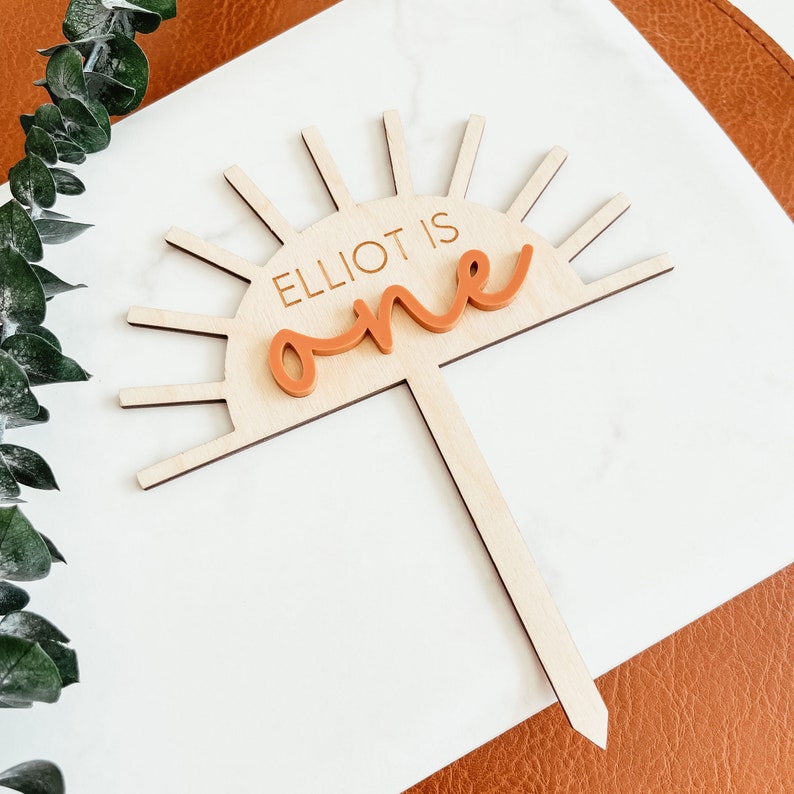 Puede incluir: Topper de pastel de madera con un dise&ntilde;o de sol y el texto "Elliot is one" en naranja. El sol tiene rayos y el texto est&aacute; en una fuente cursiva.