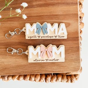 May include: Two wooden keychains with the words "MAMA" and "MOM" in white block letters. Each keychain has a bow accent, one blue and one pink. The names "Wyatt, Penelope, Theo" and "Olivia, Carter" are below the words.