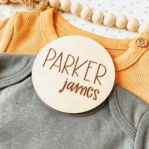 May include: A wooden round sign with the name "Parker James" engraved in brown lettering on a light brown background.