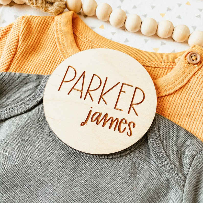 May include: A wooden round sign with the name "Parker James" engraved in brown lettering on a light brown background.