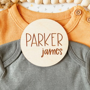 May include: A round wooden sign with the name "Parker James" engraved in brown lettering. The sign is placed on top of a gray and a yellow baby onesie.