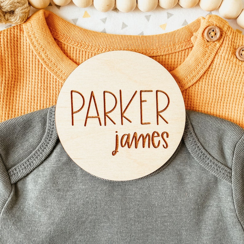 May include: A round wooden sign with the name "Parker James" engraved in brown lettering. The sign is placed on top of a gray and a yellow baby onesie.