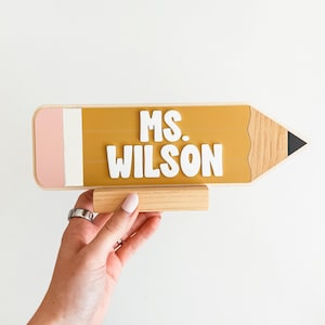 Peut inclure: Panneau en forme de crayon en bois avec le texte "MS. WILSON" en lettres blanches. Le crayon a un corps jaune, une gomme rose et une pointe noire. Il est maintenu par un support en bois.