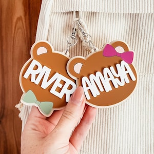 May include: Two brown bear-shaped keyrings with white text reading "RIVER" and "AMAYA". The keyring on the left has a green bow tie, and the keyring on the right has a pink bow. Both keyrings are attached to a silver clasp.