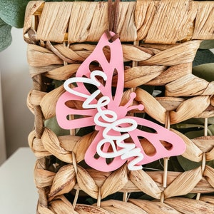 May include: Pink wooden butterfly ornament with the word "Welcome" written in white on it. The ornament is hanging from a woven basket.