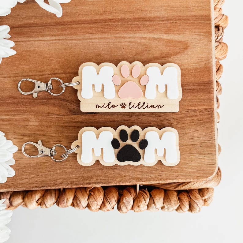 Puede incluir: Dos llaveros de madera con la palabra "MOM" en letras blancas. Uno presenta una huella de pata rosa y los nombres "milo" y "lillian". El otro tiene una huella de pata negra. Ambos tienen llaveros plateados.