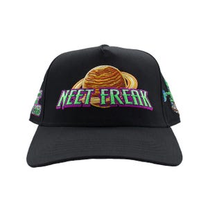 May include: A black baseball cap featuring an embroidered graphic of a planet with rings on the front. The text "NEET FREAK" is embroidered in light green with a purple outline across the centre. Additional embroidered graphics appear on the sides.