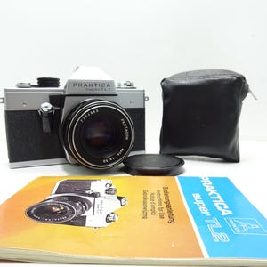 Op de afbeelding: Een Praktica Super TL2 camera met een zwart en zilverkleurig lichaam, een lens en een zwarte leren etui. De camera heeft de tekst "PRAKTICA Super TL2" aan de bovenkant. Een handleiding met de afbeelding van de camera ligt voor de camera.