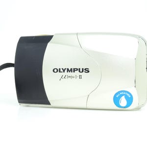 May include: A silver and black Olympus µ [mju:]-II camera with a black wrist strap. The camera has the text "OLYMPUS µ [mju:]-II" printed on the front. A blue "All Weather" sticker is on the bottom right corner.