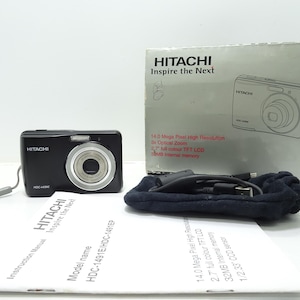 以下が含まれることがあります： 前面に「HITACHI」の文字が書かれた、銀色のレンズを持つ黒いデジタルカメラ。カメラは白い表面に置かれ、隣には「HITACHI Inspire the Next」と「モデル名 HDC-1491E/HDC-1491EP」の文字が書かれた白い取扱説明書がある。