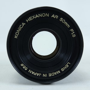 May include: A black Konica Hexanon AR 50mm F1.8 camera lens. The lens has a 55mm diameter and is made in Japan. The lens is circular with a clear glass center.