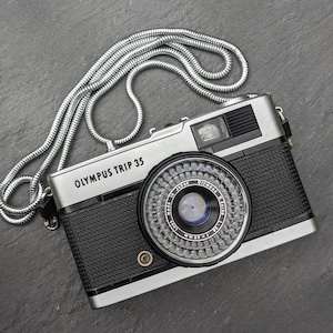 Op de afbeelding: Een Olympus Trip 35 camera met een zilveren behuizing en zwarte textuur grip. De camera heeft een lens met de tekst "Olympus D.Zuiko 40mm" en een zilveren kettingriem. Op een donkergrijze ondergrond.