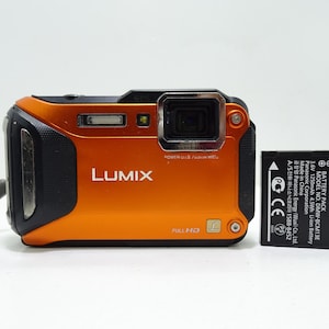 May include: Orange and black Lumix digital camera with a Full HD label. A black Panasonic battery pack is next to the camera. The camera has a lens, flash, and various buttons.