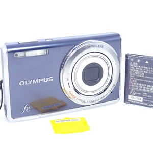 Puede incluir: Una cámara digital Olympus plateada y azul con una lente que dice "4x Wide Optical Zoom". La cámara va acompañada de una batería y una tarjeta de memoria amarilla. La cámara tiene el texto "fe" en la parte delantera.
