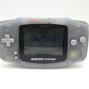 Könnte beinhalten: Ein transparent grauer Game Boy Advance Handheld-Konsolen. Der Bildschirm zeigt "GAME BOY" und "GAME BOY ADVANCE" in weißem Text an. Es verfügt über ein Steuerkreuz, Tasten und einen Lautsprecher.