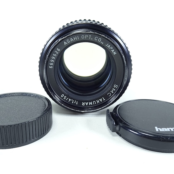 以下が含まれることがあります： 「SMC TAKUMAR 1:1.4/50」と「ASAHI OPT. CO., JAPAN」の文字が印刷された黒いカメラレンズ。レンズには黒いレンズキャップが2つ付属しており、1つには「hama.」の文字が印刷されています。