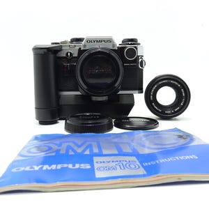 Puede incluir: Una cámara de película Olympus OM-10 negra y plateada con un objetivo. La cámara está sobre una superficie blanca con dos tapas de objetivo y un manual de instrucciones azul. El manual tiene el texto "Olympus OM-10 Instructions" impreso.