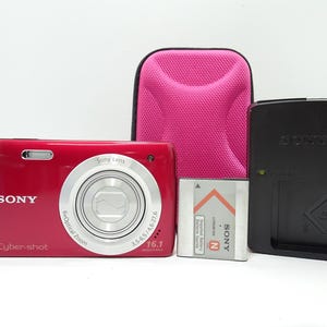 May include: A red Sony Cyber-shot digital camera with a silver lens, a black charger, a pink case, and a battery. The camera has 6x optical zoom and 16.1 megapixels.
