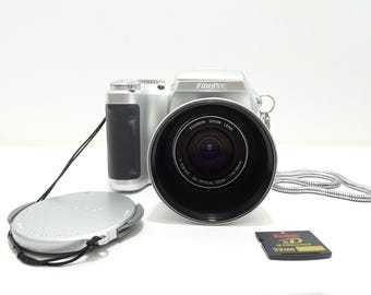 Pode incluir: Câmera digital FinePix prateada com uma lente grande, tampa da lente e um cartão de memória Kodak XD de 32 MB. A câmera tem uma empunhadura preta e uma alça prateada. A lente é rotulada como "FUJINON ZOOM LENS".